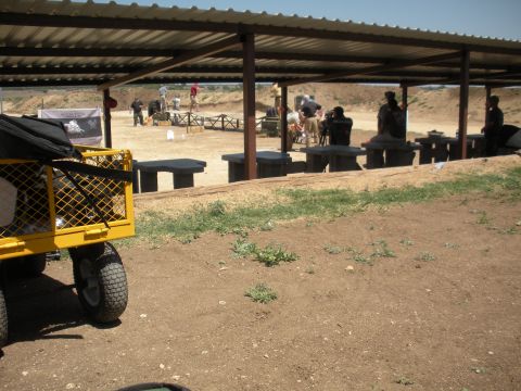 1996 Ford Bronco - A covered area at a shooting range provides shade for people and tables, with a yellow utility cart visible in the foreground.