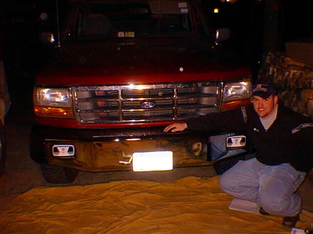Man crouching by red Ford Bronco front end at night, black bumper and chrome grille visible