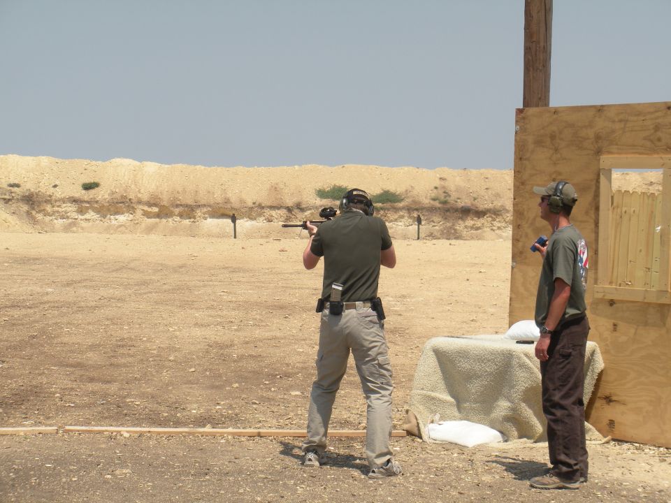 Two men at an outdoor shooting range, one aiming a rifle