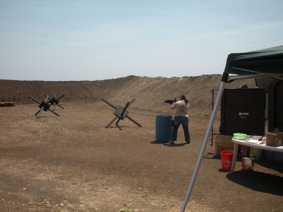 Shooting range with a person aiming a rifle, blue barrel, and anti-tank obstacles under a clear sky