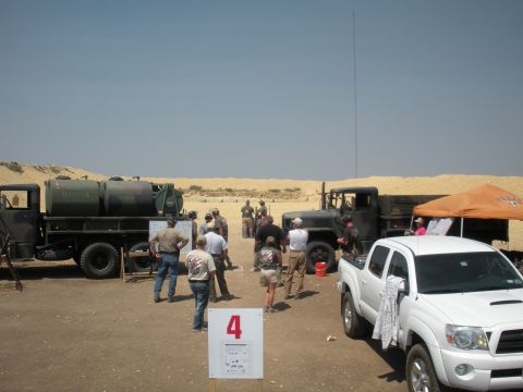1996 Ford Bronco - A group of people gather around military trucks and a white pickup truck at a desert shooting range, with a sign numbered '4' in the foregr…