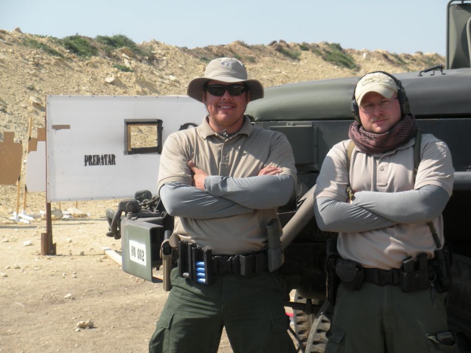 Two men in tactical gear stand in front of a military-style vehicle and a shooting target board. Text 'PREDATAR' is visible on the board