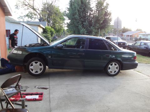 2003 Ford Taurus - Mechanic works on a green sedan parked in a driveway with the hood up.