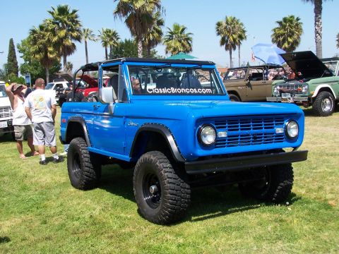 1990 Ford Bronco - Blue Ford Bronco at an outdoor car show with other vehicles and palm trees.