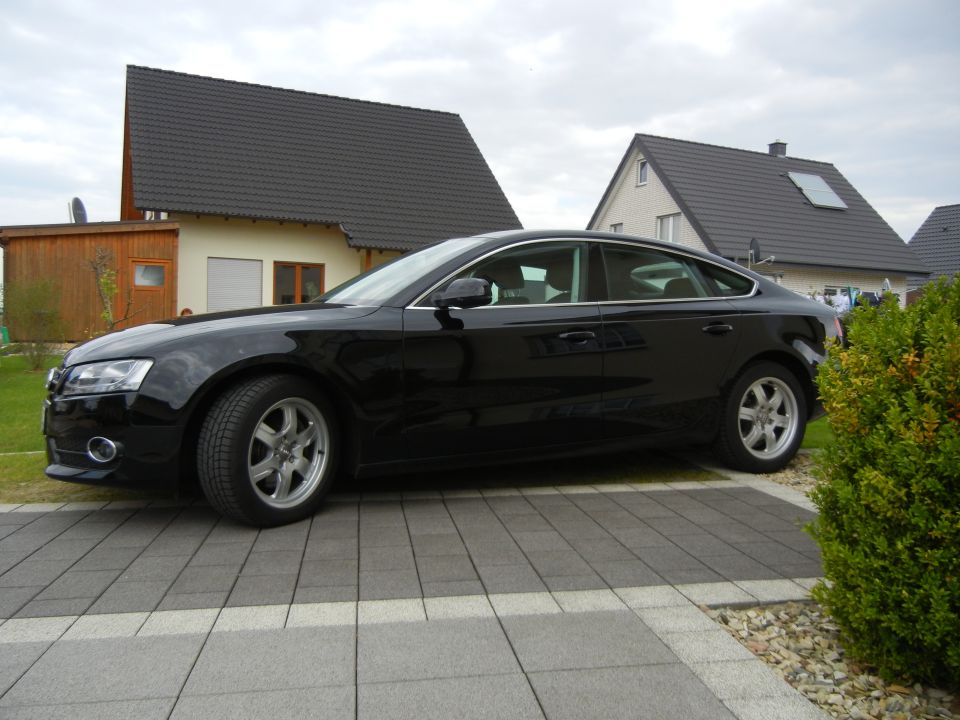 Black Audi sedan parked on a paved driveway in front of houses