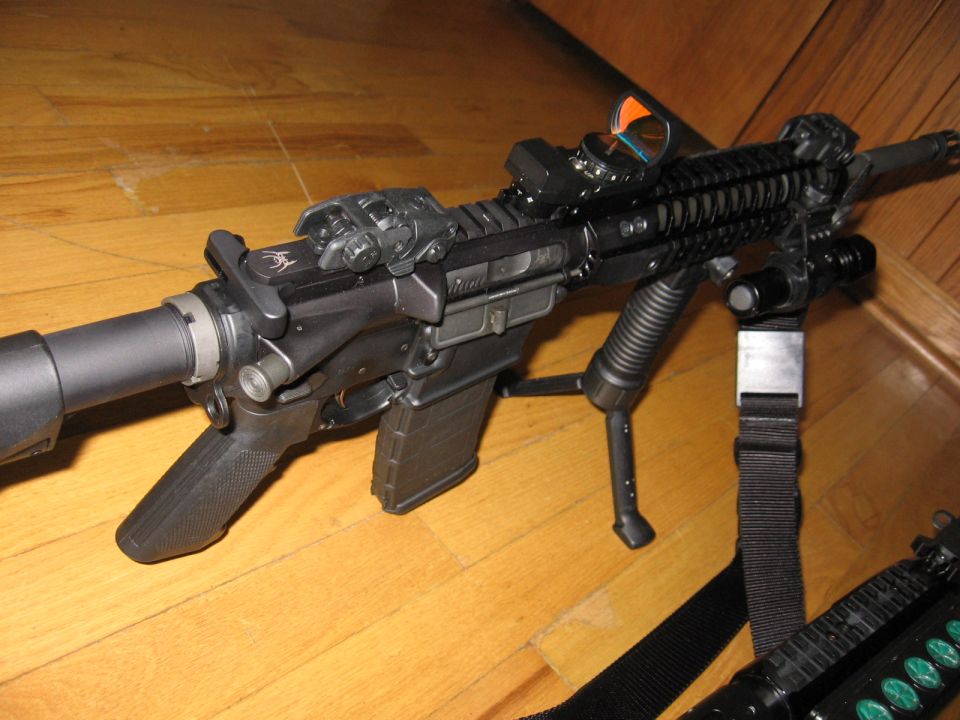 Close-up of a black AR-pattern rifle with optic and bipod on a hardwood floor