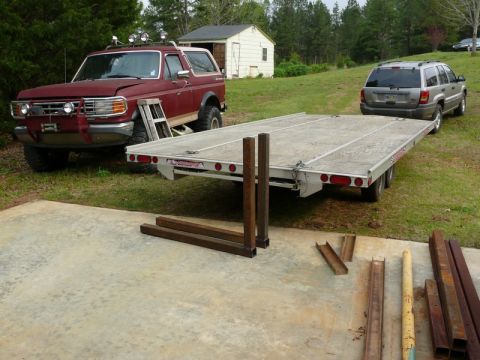 1976 Chevrolet Corvette - A red Ford Bronco and a silver SUV are parked near a flatbed trailer with metal pieces on the ground.