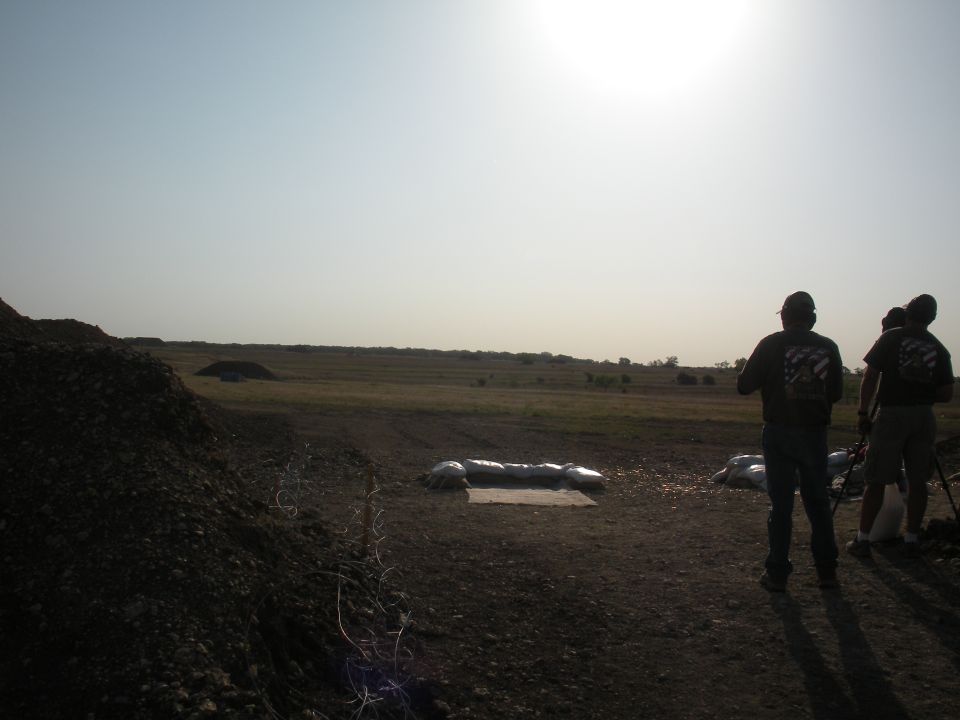 Three people stand in a dusty field under a bright sky, with a pile of dark gravel to the left