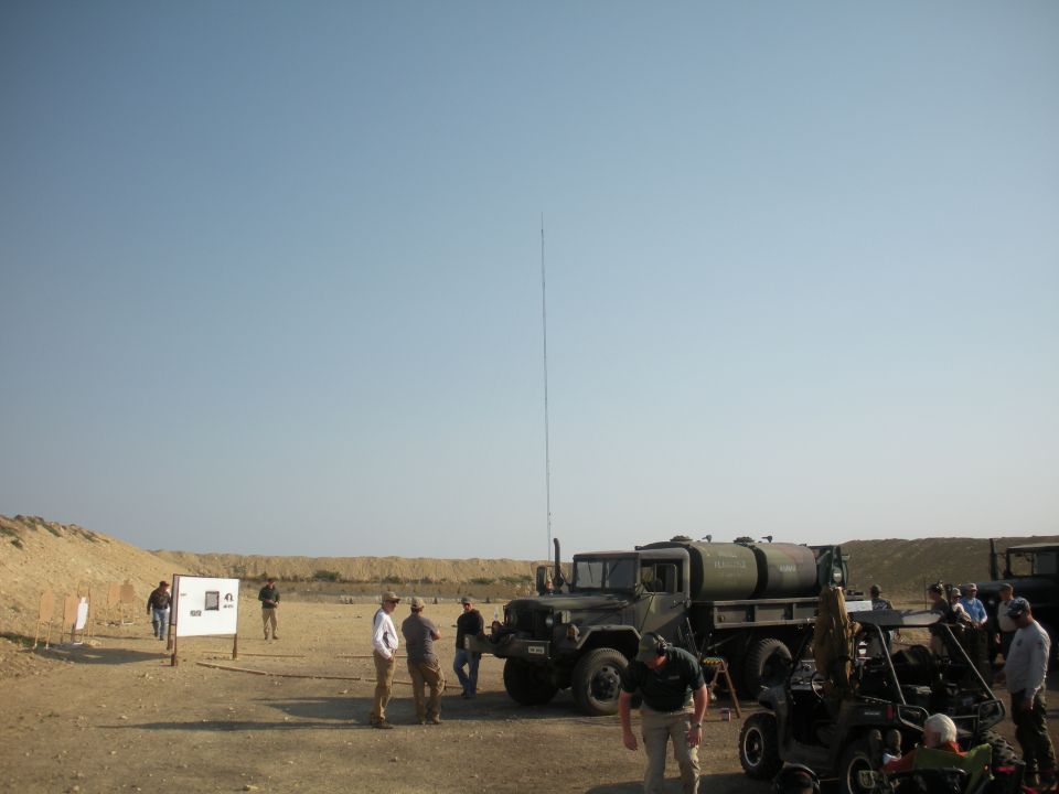 Military truck with water tank and tall antenna parked at a shooting range with people around