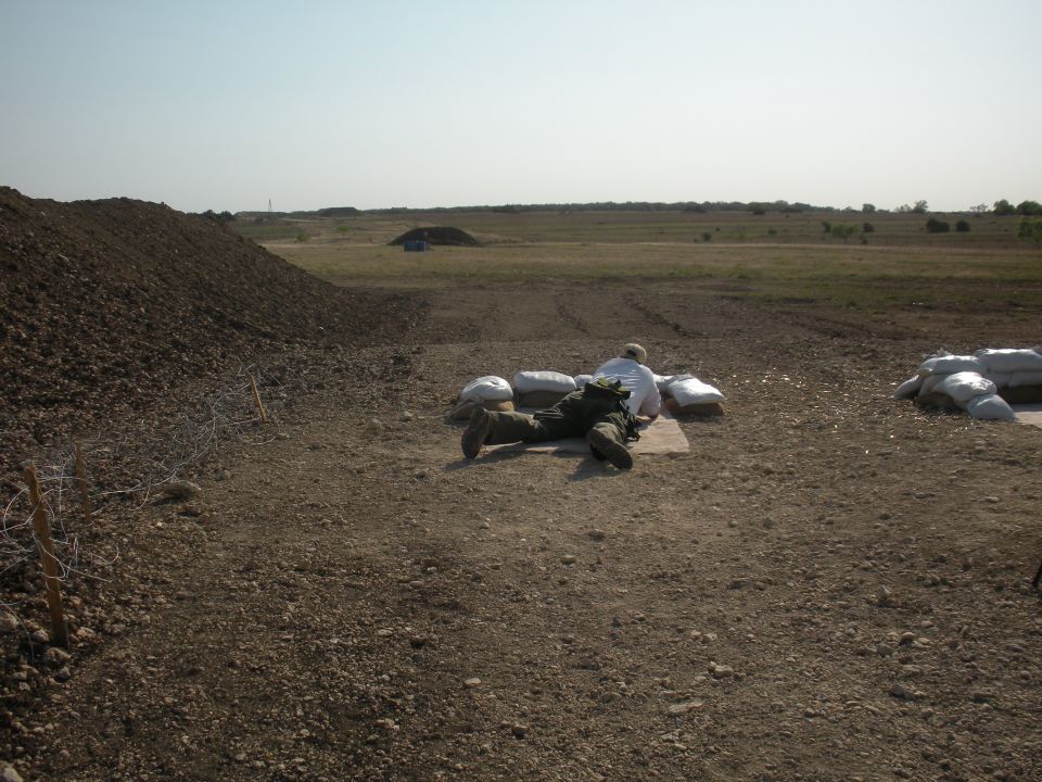Person in camouflage lying prone behind sandbags in a dry, open field