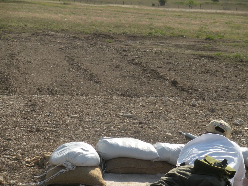 Person aiming rifle from behind sandbags in a dry, grassy field