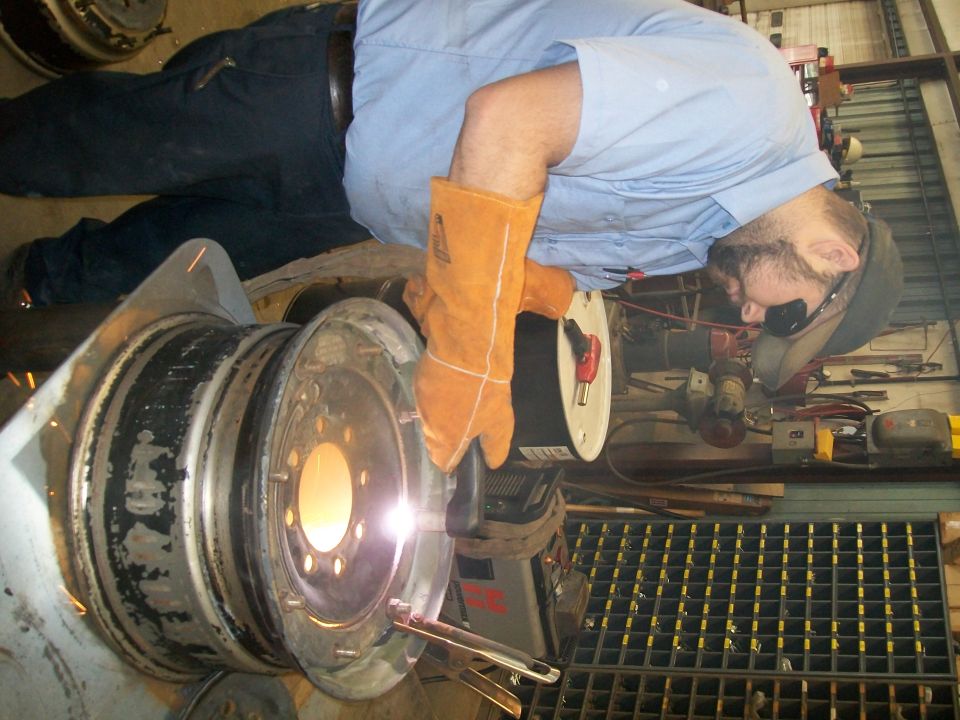 Welder in orange gloves and safety glasses sparks metal on a large wheel rim in a workshop