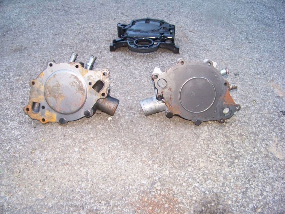 Three automotive engine parts on asphalt: two water pumps and a black timing cover