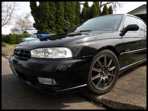 1998 Subaru Legacy - A black 1998 Subaru Legacy sedan is parked on a residential street, showing its front quarter panel and wheel.