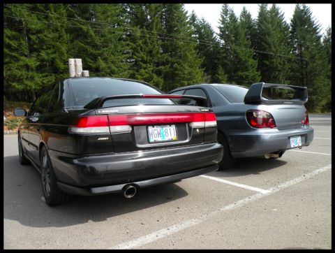 1998 Subaru Legacy - Two Subaru sedans, a black 1998 Legacy and a gray WRX STI, are parked side-by-side in a lot.