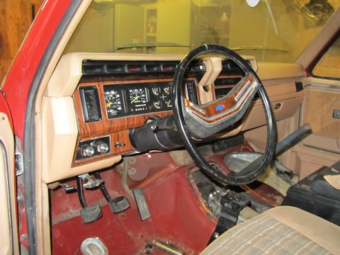 1984 Ford Bronco - The interior of a red vehicle shows a dashboard with wood-grain accents and a black steering wheel.