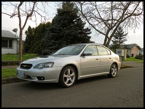 2006 Subaru Legacy - A silver sedan is parked on the side of a street in front of houses and trees.