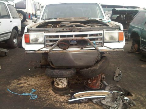 1986 Ford Bronco - A white Ford Bronco sits in a junkyard with its hood open and missing tires.