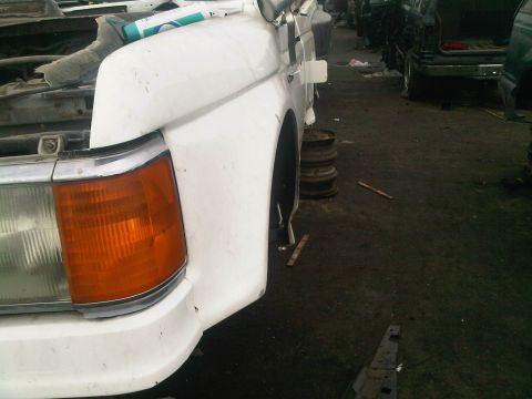 1986 Ford Bronco - Front corner of a white pickup truck in a junkyard setting with other vehicles around.