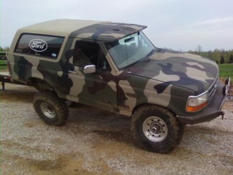 1995 Ford Bronco - Camouflaged 1995 Ford Bronco parked on a gravel surface with trees in the background.