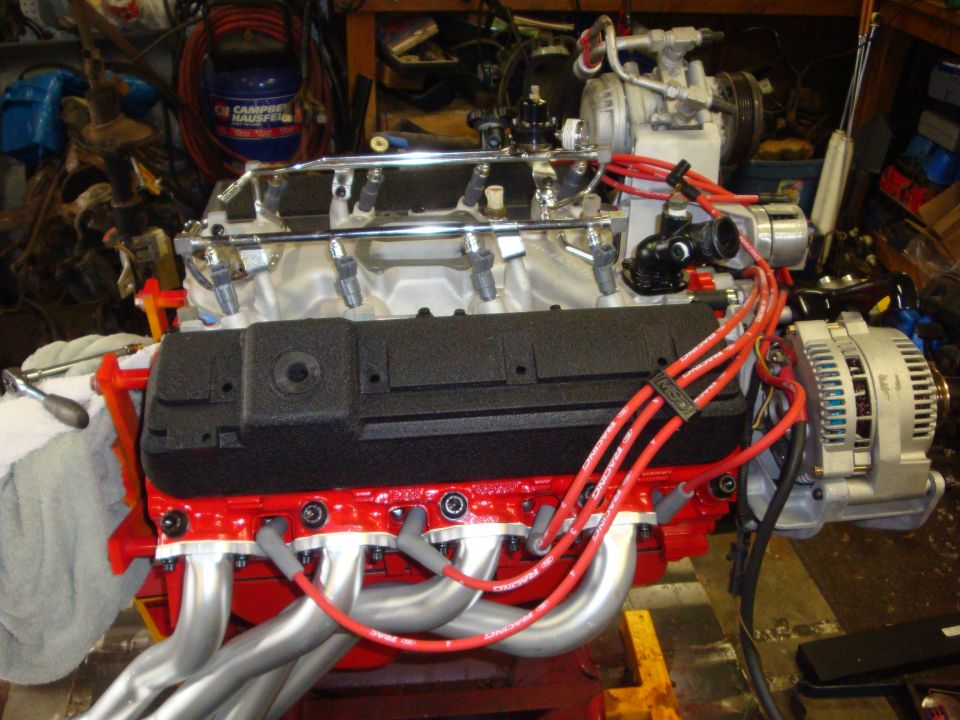 V8 engine block painted red, with silver headers, black intake manifold, and red spark plug wires. Alternator visible on the right
