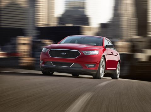 1993 Ford Taurus - A red Ford sedan is captured in motion on a highway, with a blurred cityscape visible behind it.