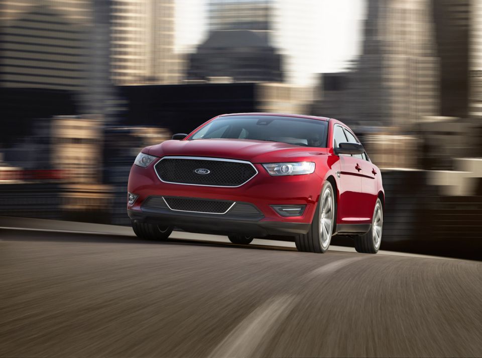 red Ford sedan driving on a road with city buildings blurred in the background
