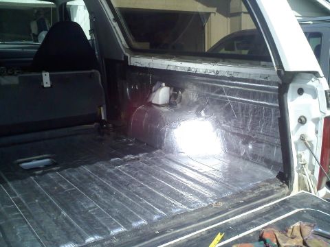 1994 Ford Bronco - Passenger-side rear cargo area of a white full-size SUV with foil-faced sound-deadening mat rolled over the floor, wheel well and inner qua…
