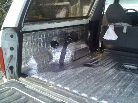 1994 Ford Bronco - Rear cargo area of a white full-size SUV mid-project, with foil-faced sound-deadening mat applied over the bare floor, rear wheel well and…