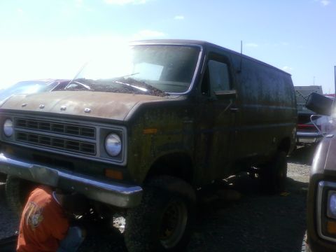 1989 Ford Bronco - A person works on the undercarriage of a rusty Ford van parked in a lot.