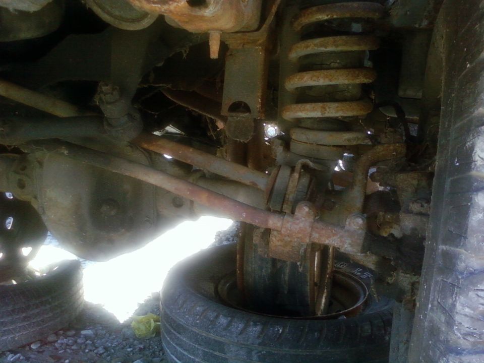 Close-up of rusted vehicle suspension parts, including a coil spring, axle, and steering linkage, with tires visible