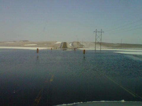 2011 Ford 427 - A flooded road creates a reflective surface, with yellow striped markers and power lines visible in the distance under a pale sky.