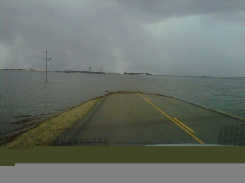 Road submerged in floodwater under a cloudy sky. Power lines visible in the distance