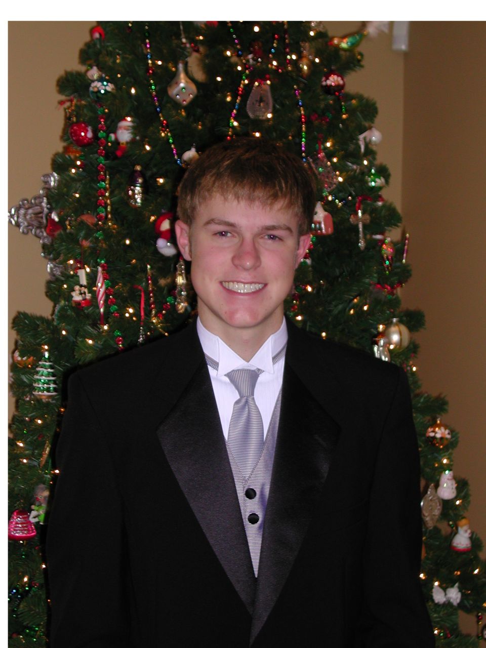 Young man with braces in a black tuxedo and silver tie in front of a Christmas tree