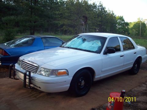 1998 Ford Crown Victoria - Front three-quarter view of a white Ford Crown Victoria sedan on red clay ground, fitted with a black tubular push bar on the front bumper…