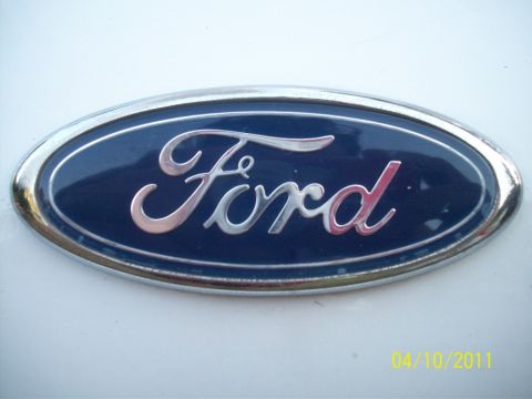 1998 Ford Crown Victoria - Extreme close-up of a blue Ford oval badge with chrome script and chrome surround mounted on a white painted body panel. The camera date st…