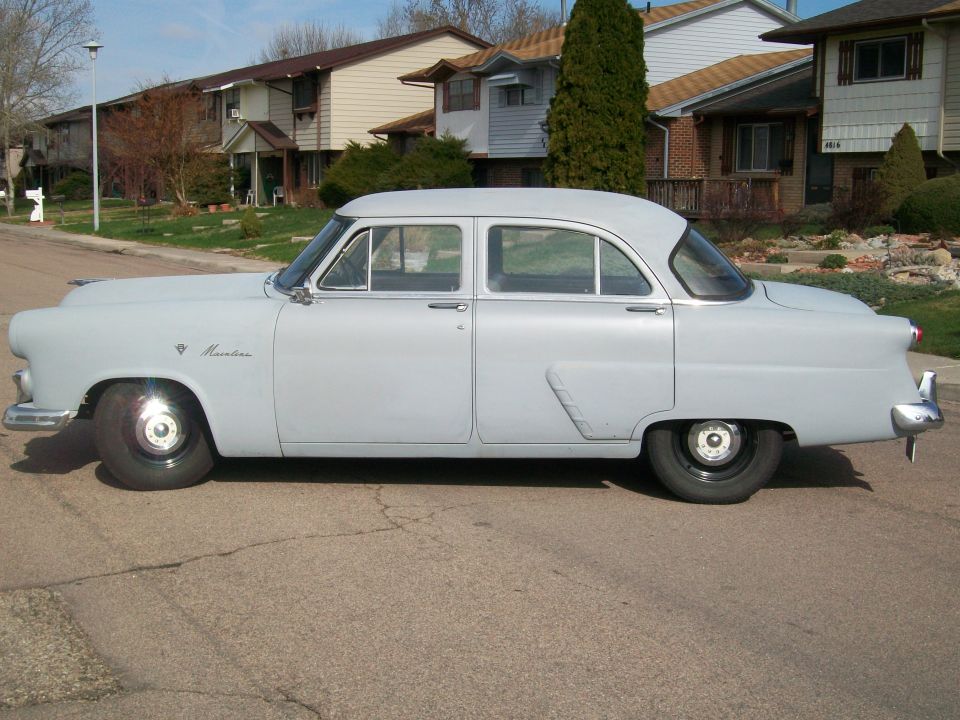 Light gray 1950s Ford Mainline four-door sedan in side profile on a residential street.