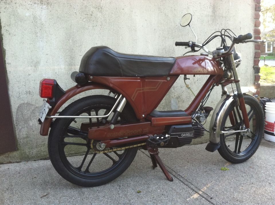 Side view of a brown Garelli moped parked next to a wall. It has a black seat and black wheels