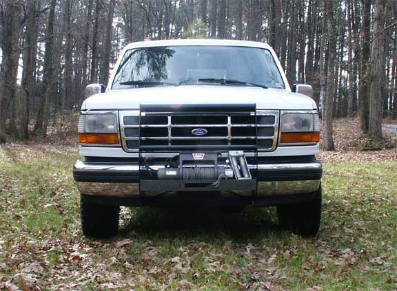 Front view of white Ford Bronco with black grille guard and front winch in a wooded yard