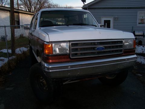 1989 Ford Bronco - Three-quarter front view of a lifted two-tone brown-and-white Ford Bronco parked on a driveway beside a gray house, wearing a mismatched wh…