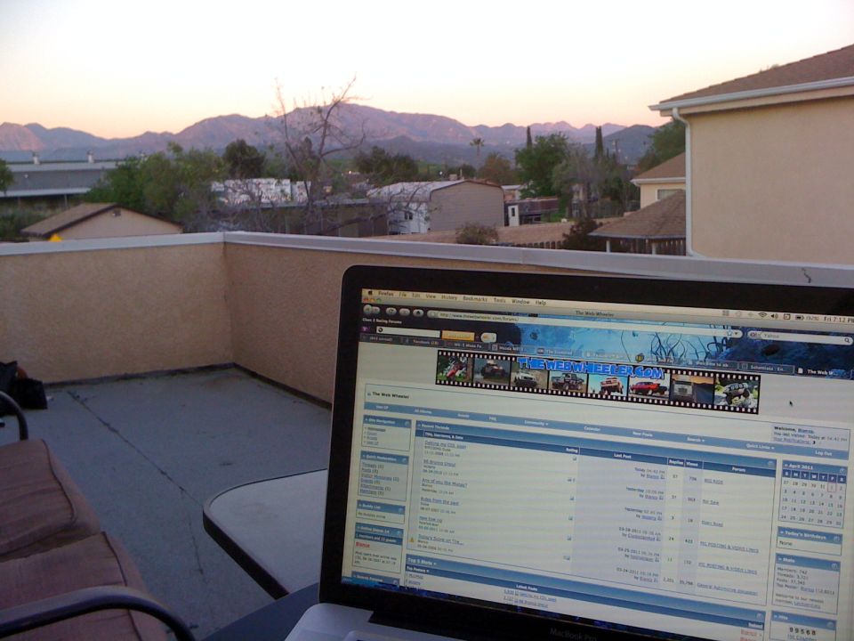 Laptop on patio table, showing a website with car photos, with mountains in background at sunset