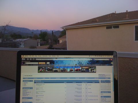 1968 Ford Mustang - A laptop displays a forum page with a banner various vehicles, set against a backdrop of mountains and a house at dusk.
