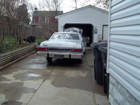 1970 Ford Ranchero - A silver sedan with a chrome bumper and horizontal taillights is parked in a driveway next to a garage.