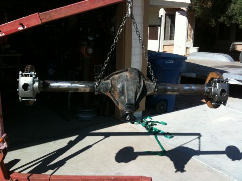 2009 Suzuki Samurai - A rusty rear axle assembly with disc brakes is suspended by chains, with a trailer and trash cans visible in the background.