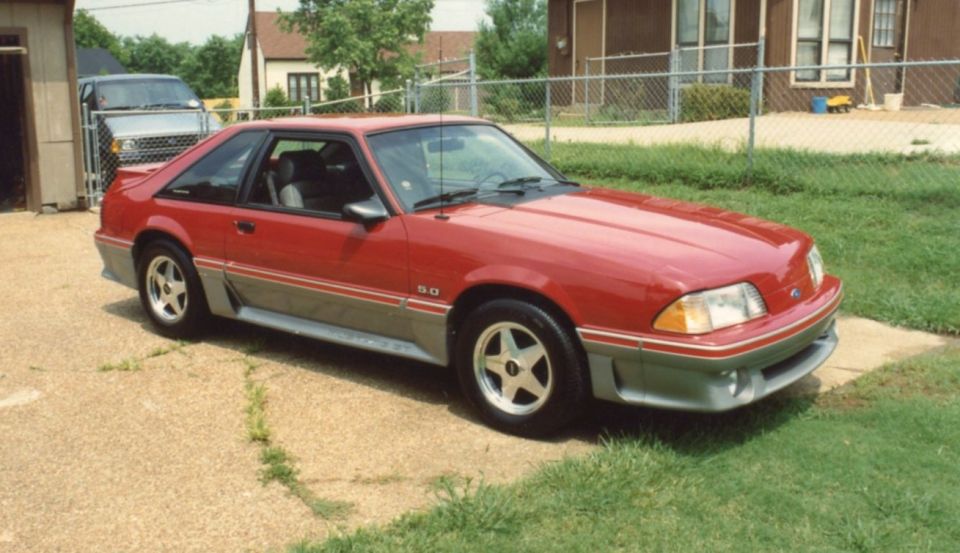 Red 1987 Ford Mustang GT coupe with gray lower body cladding and a 5.0 badge, parked on a gravel driveway