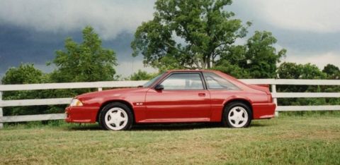 2007 Ford Mustang - This red Ford Mustang coupe, likely from the late 1980s, is shown in profile next to a white fence.