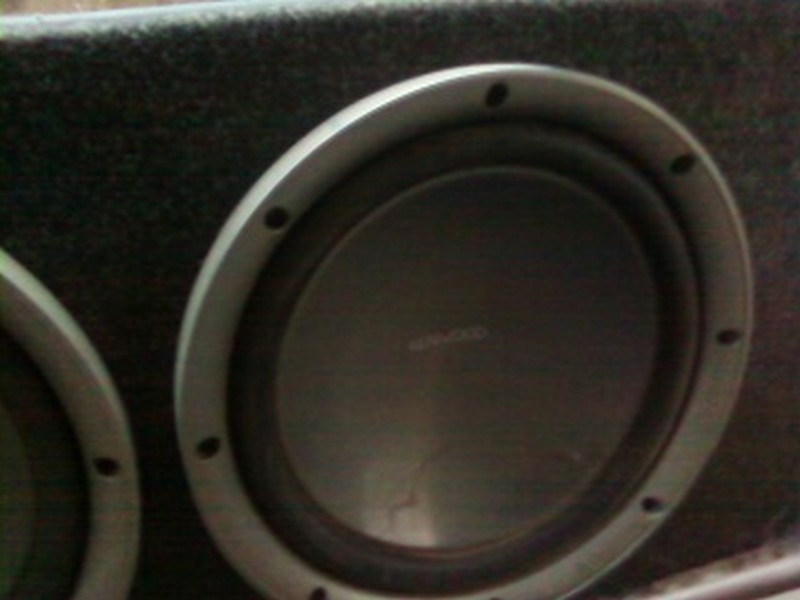 Close-up of a car subwoofer in a box. Text on the speaker reads 'KENWOOD'