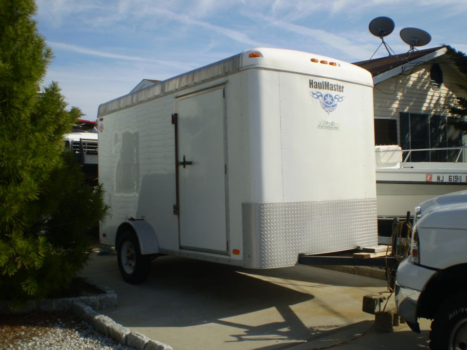 White enclosed cargo trailer with diamond plate lower front, parked next to a boat and part of a truck