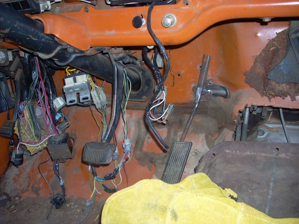 Vehicle interior showing exposed wiring, pedals, and orange dashboard. A yellow cover is visible