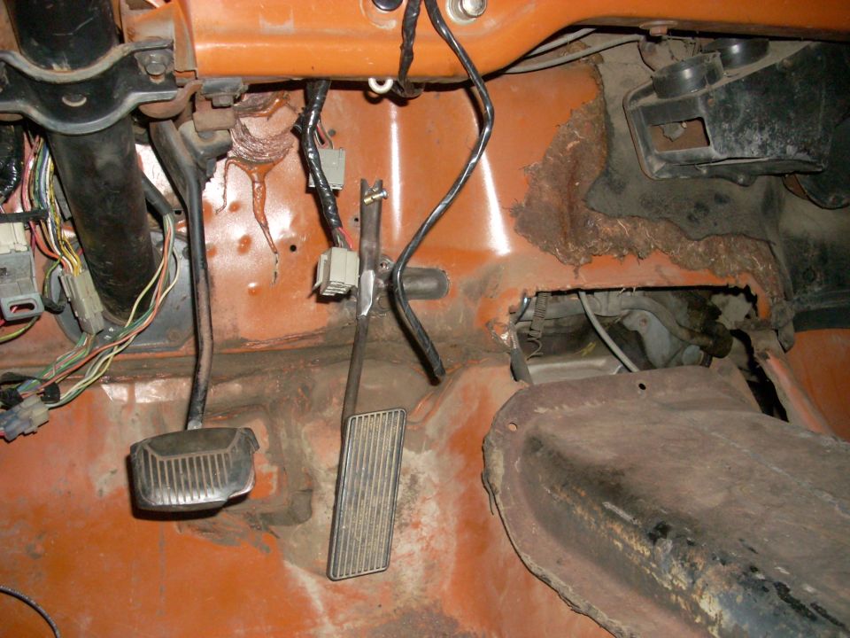 Interior floor of a vehicle showing brake and accelerator pedals, exposed wiring, and rust